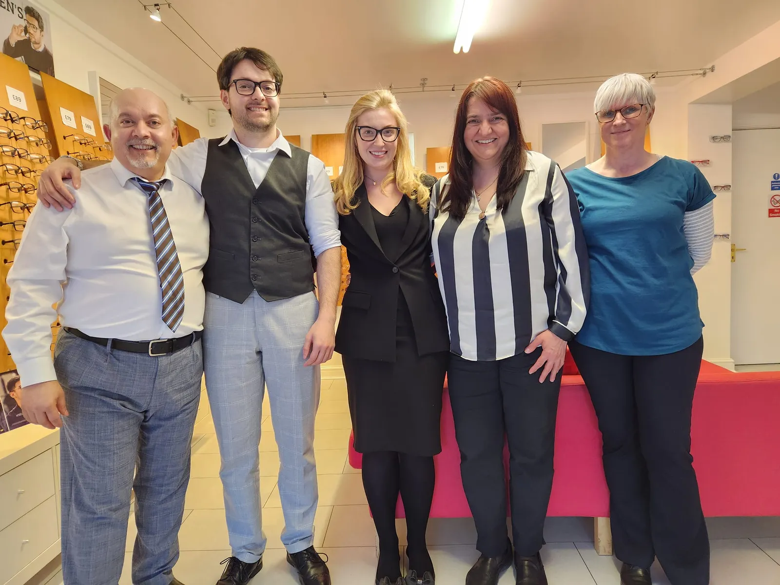The Mable Opticians team smiling together in front of the frame wall at the practice in Mablethorpe